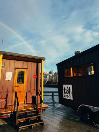 Two tiny house cabins on a waterfront dock with wooden steps, an urban skyline in the background and a faint rainbow arcing across a blue sky.