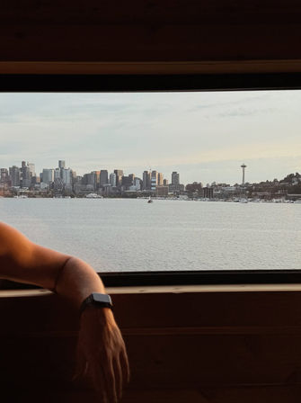 Window view of the Seattle skyline with the Space Needle across a calm waterfront, foreground shows a relaxed arm wearing a smartwatch resting on a wooden ledge at dusk.