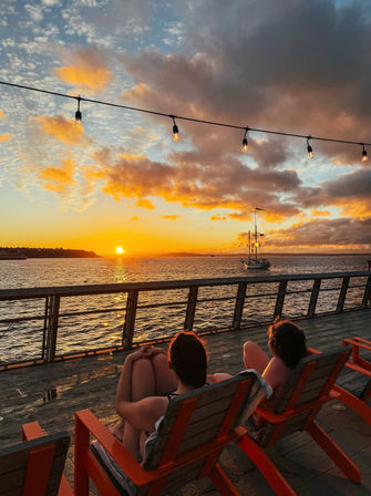 Two people relaxing in orange chairs on a waterfront pier at sunset, string lights overhead and a sailboat on the glowing sea