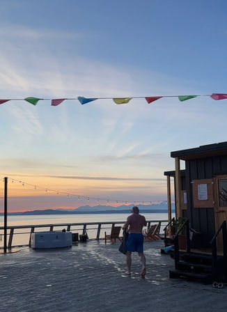 Sunset over a waterfront wooden deck/pier with string lights and colorful pennant flags, Adirondack chairs and a hot tub, calm water and distant mountain silhouette, person in swim shorts walking toward the railing.