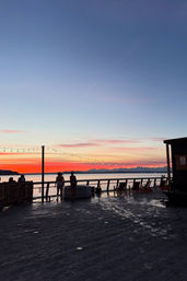 Silhouetted people on a waterfront deck strung with lights, gazing over calm water as a vibrant orange-pink sunset fades to deep blue above distant mountains.