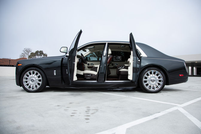 Black luxury sedan with rear-hinged coach doors open showing cream leather interior and polished alloy wheels, parked in an empty rooftop parking lot under an overcast sky