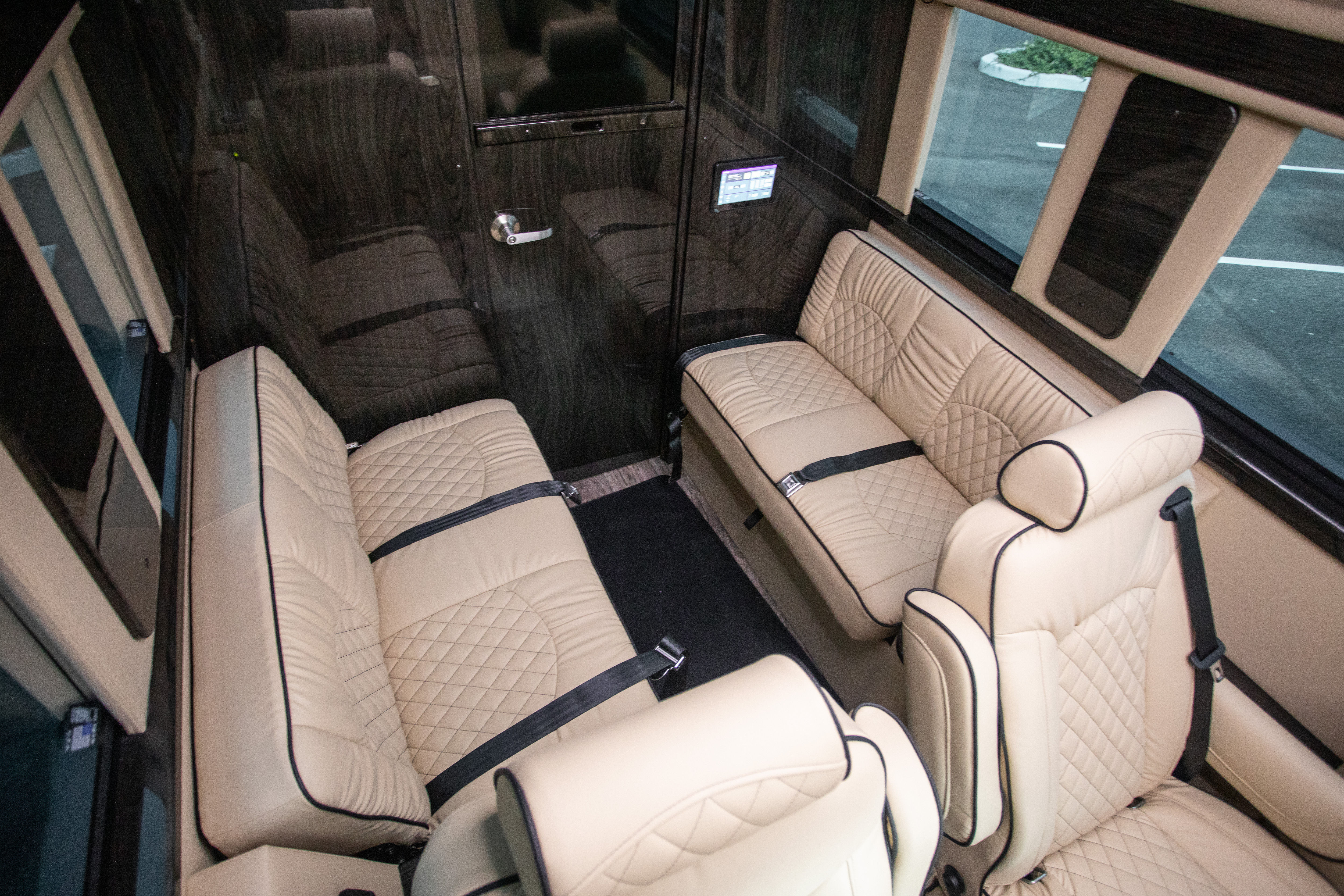 Luxury executive van interior with beige quilted leather seats and black piping, two bench seats facing each other plus captain chairs, visible seat belts, dark wood-paneled walls and windows showing a parking lot — upscale shuttle-style passenger cabin.