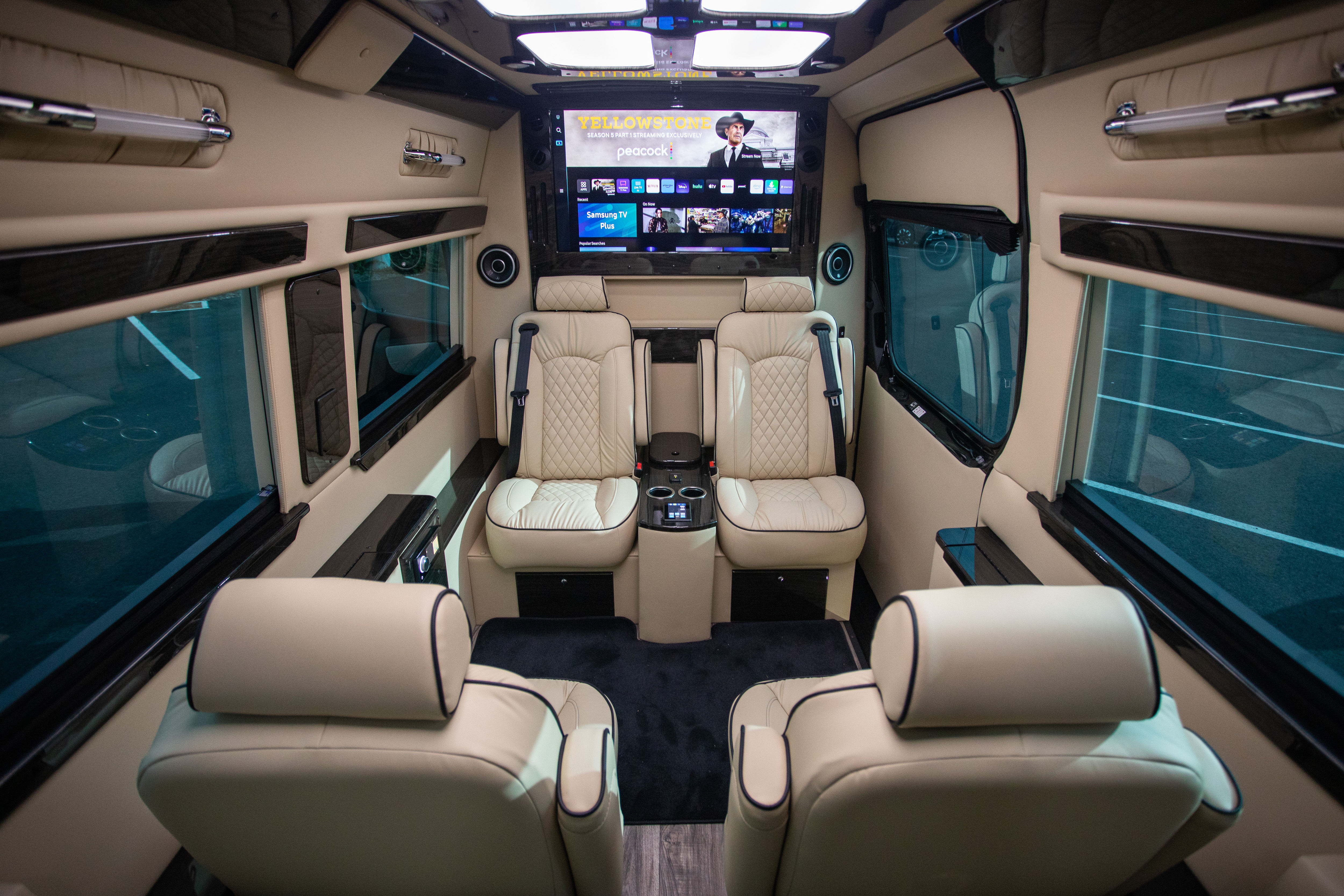 Interior of a luxury executive shuttle van with four cream quilted leather captain seats, center console with cupholders, widescreen entertainment monitor, ambient LED lighting and tinted windows