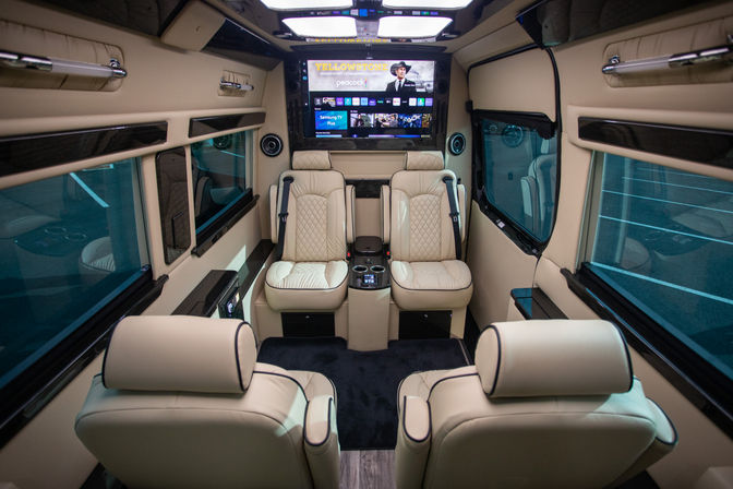 Interior of a luxury executive shuttle van with four cream quilted leather captain seats, center console with cupholders, widescreen entertainment monitor, ambient LED lighting and tinted windows