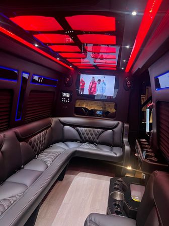 Luxury party bus interior with red LED ceiling panels, quilted black leather wraparound seating, cupholders, wood-look floor and a wall-mounted TV — upscale nightlife transportation vibe.