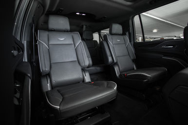 Spacious luxury SUV interior with two black leather captain seats featuring white piping and headrests, third-row bench visible behind, and a window view of an indoor parking garage.