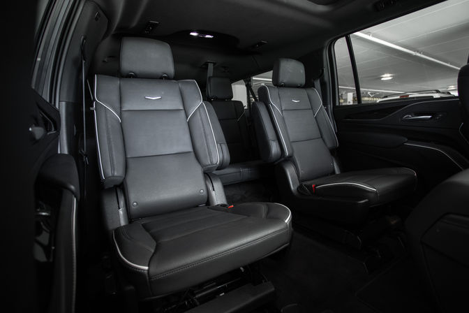 Spacious luxury SUV interior with two black leather captain seats featuring white piping and headrests, third-row bench visible behind, and a window view of an indoor parking garage.