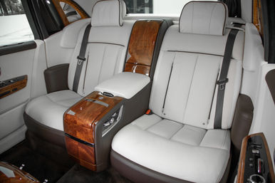 Luxury car rear cabin with white leather seats, burl wood center console and armrest, black seatbelts and plush dark carpet
