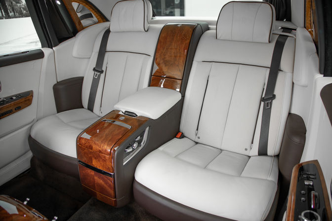 Luxury car rear cabin with white leather seats, burl wood center console and armrest, black seatbelts and plush dark carpet
