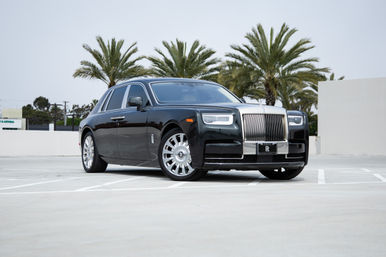 Glossy black luxury sedan with a gleaming chrome grille and polished wheels, parked on a rooftop lot framed by palm trees.