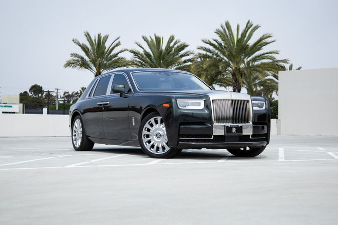 Glossy black luxury sedan with a gleaming chrome grille and polished wheels, parked on a rooftop lot framed by palm trees.