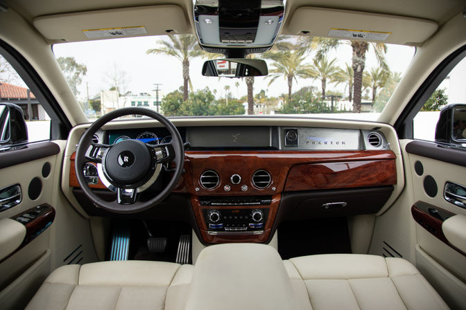 Luxury car interior with cream leather seats, glossy wood dashboard, chrome-trim steering wheel and analog clock, palm trees visible through the windshield