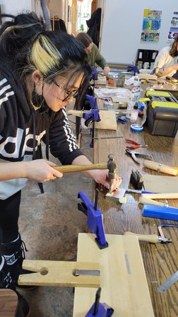 Focused person hammering a metal stamp on a small metal plate at a cluttered wooden workbench in a hands-on craft workshop with clamps, pliers and toolboxes.
