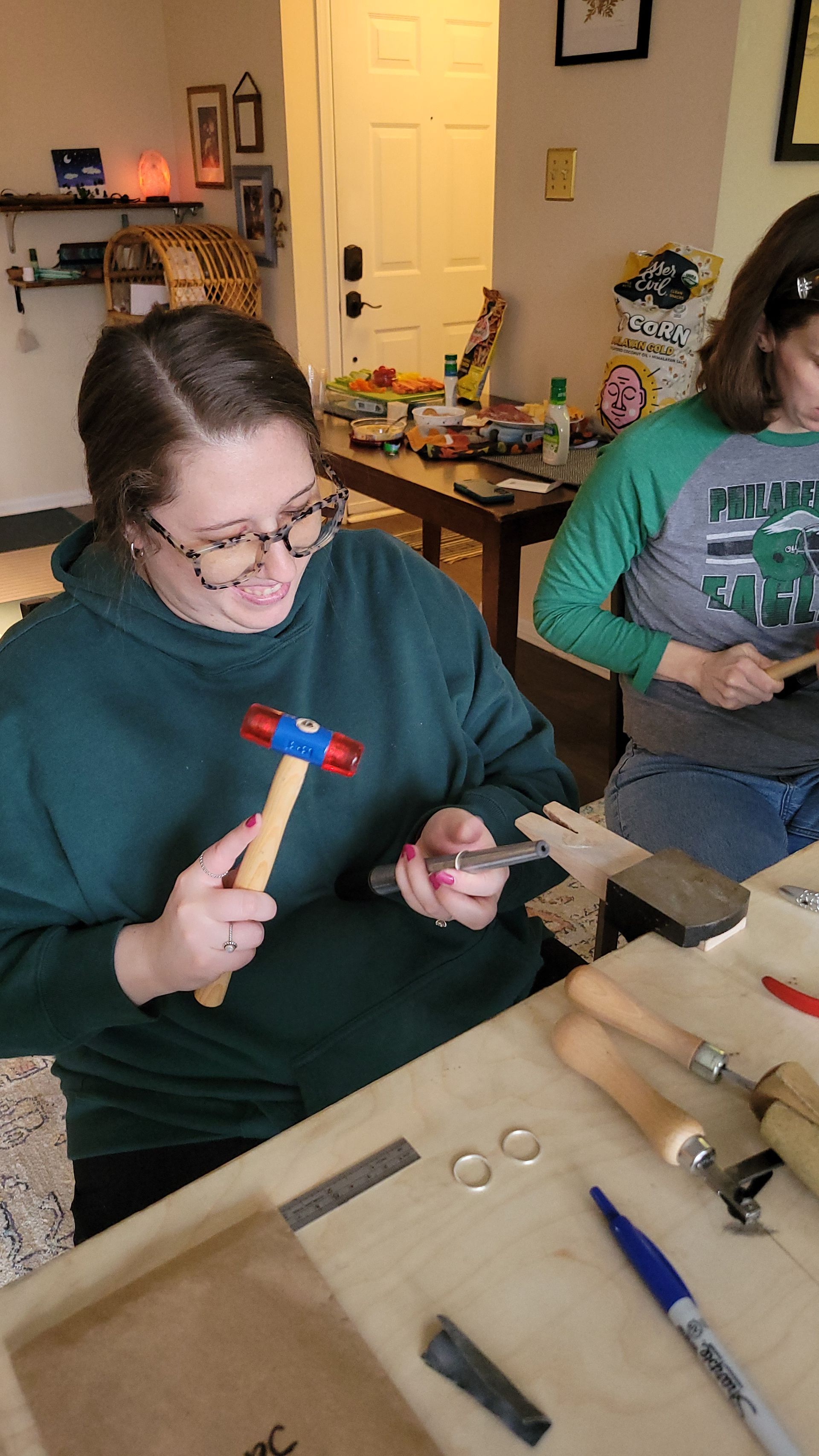 Cozy living-room craft session making rings, person holding a red-and-blue jeweler’s mallet and mandrel over an anvil, rings and tools on the table and snacks in the background.