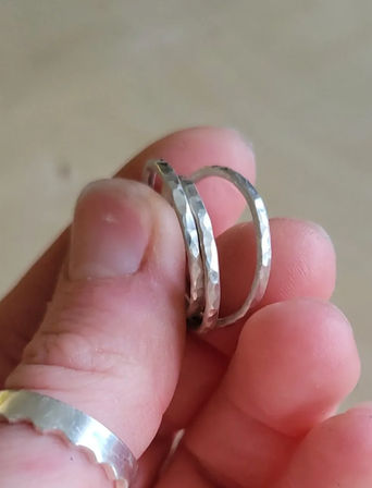 Close-up jewelry shot of a hand holding two thin hammered silver-tone stacking rings with a textured faceted finish, scalloped silver band on the thumb, neutral beige background.