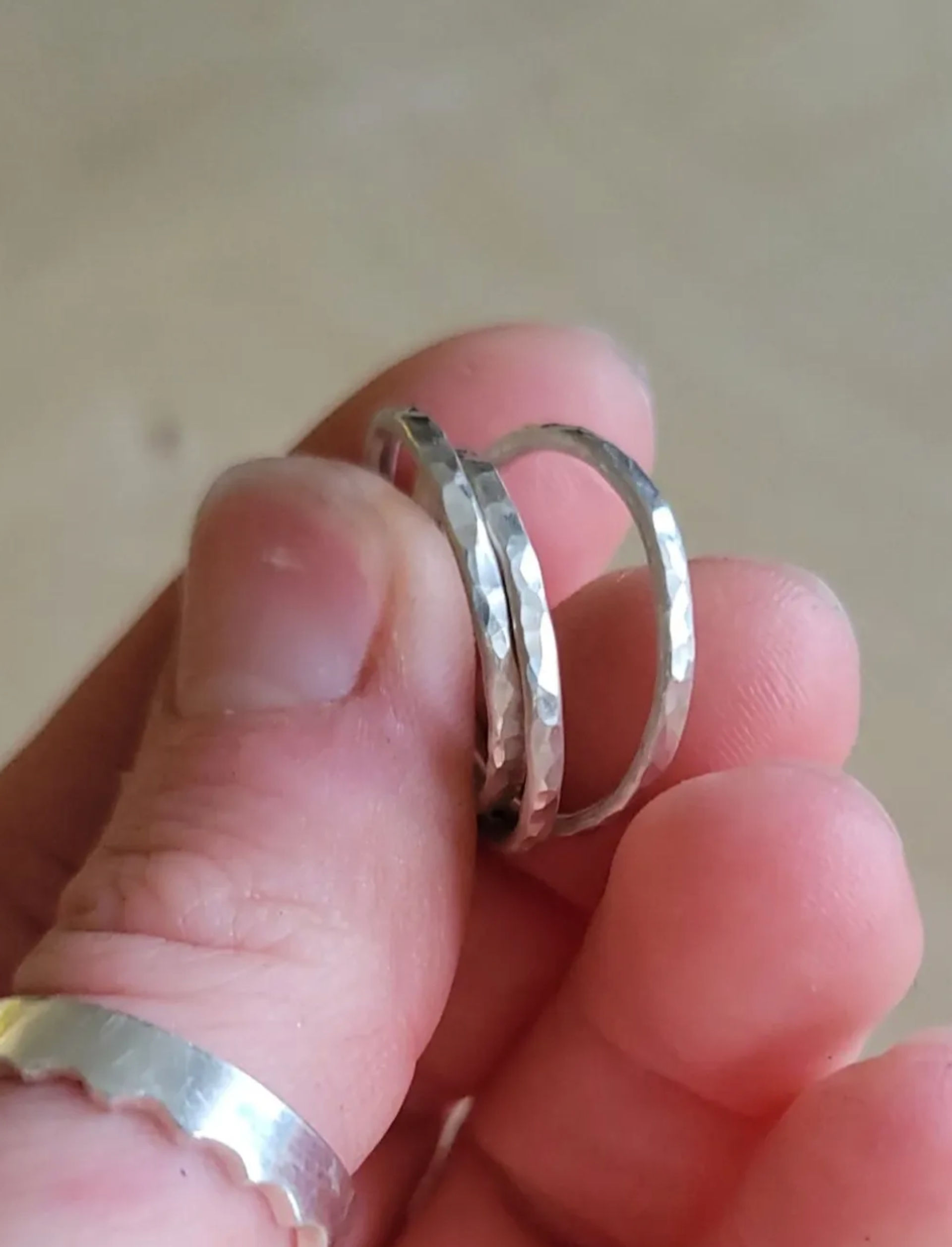 Close-up of hand holding two hammered silver stacking rings with textured facets against a neutral background