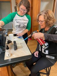 Two women at a home workbench soldering jewelry with a handheld torch over a small anvil and firebrick — indoor metalsmithing craft session