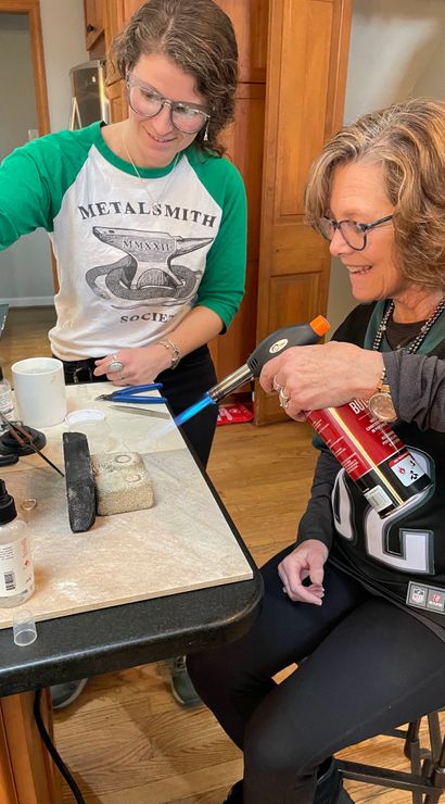 Two women at a home workshop bench practicing metalsmithing — one uses a butane torch to solder a ring on a firebrick while the other assists with tools and supplies.
