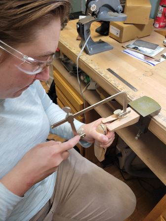 Close-up of person wearing safety glasses using a jeweler's saw on a small piece held in a wooden bench pin at a busy craft workbench