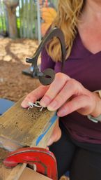 Close-up of hands using a jeweler's saw to cut metal wire on a wooden bench pin clamped with a red C-clamp at an outdoor jewelry-making station