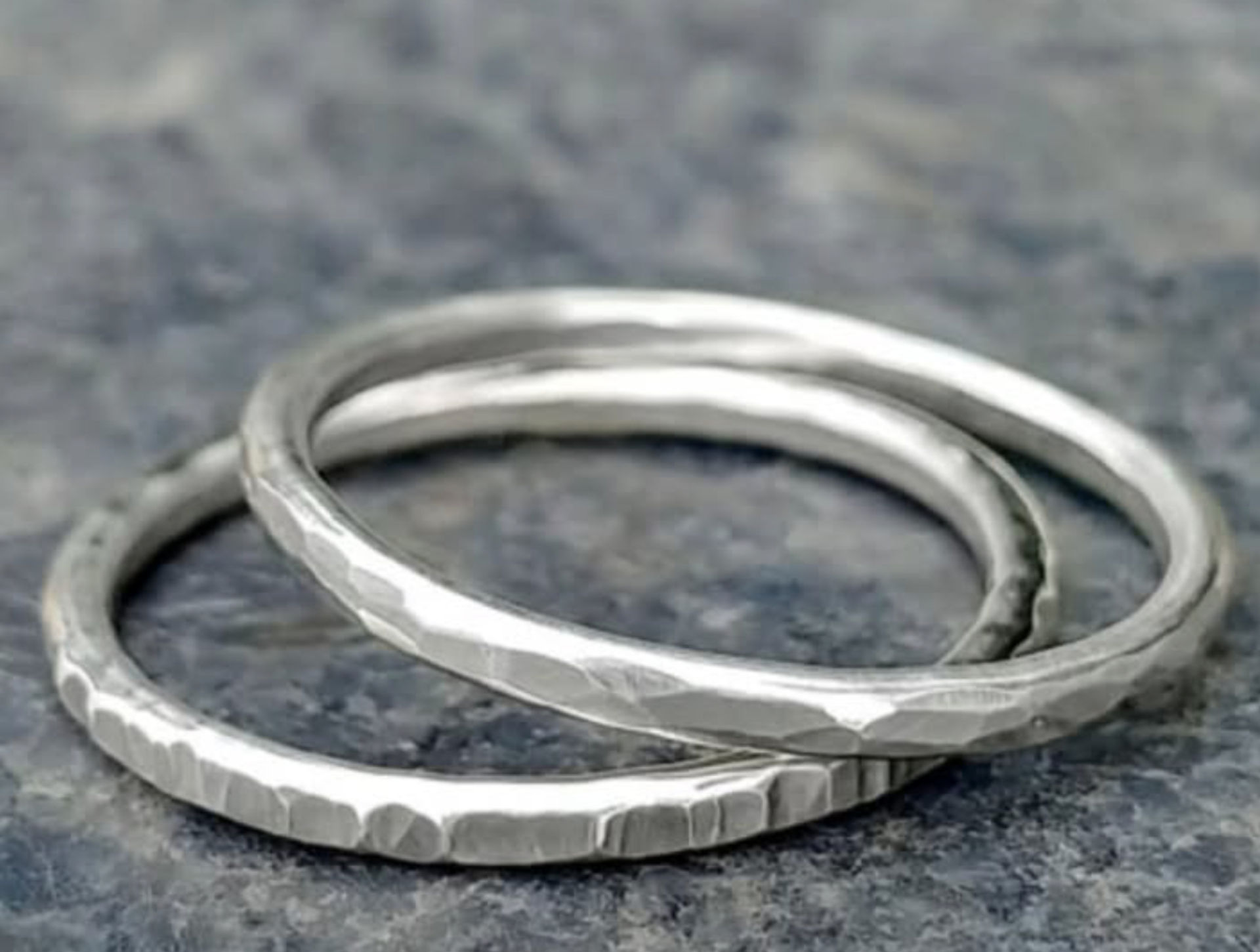 Close-up of two hammered silver stacking rings with subtle facets and rustic matte finish arranged on a textured gray slate surface