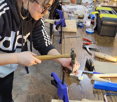 Participant metal-stamping a small plate on a wooden workbench in a busy craft studio, surrounded by clamps, hammers, pliers and toolboxes.