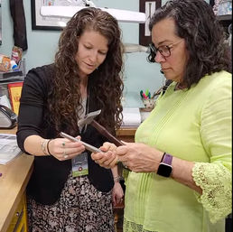 Two women at a craft studio workbench sharing tools, focused on a metal rod and pliers during a hands-on jewelry-making demonstration.