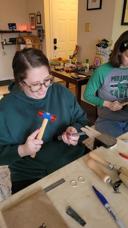 Cozy home craft workshop: person in a green hoodie uses a small mallet and ring mandrel to shape metal rings at a workbench, another person works beside them, with rings, hand tools, a marker and snacks on the table.