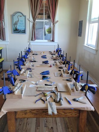 Sunlit craft studio with a long wooden table set up for a jewelry-making class — multiple bench stations with blue clamps, pliers, hammers, wire and hand tools, striped curtains and windows in the background.
