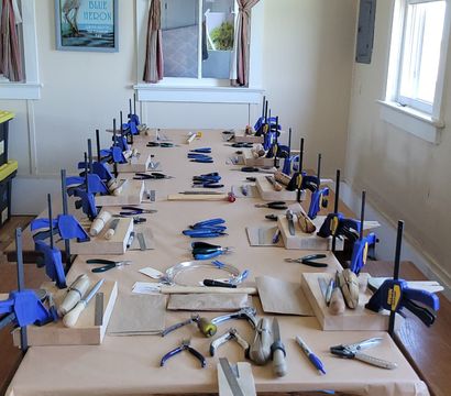 Long wooden table set up for a group jewelry-making workshop with individual bench blocks, blue clamps, pliers, hammers, files and wire coils arranged at each station in a bright room with windows and striped curtains