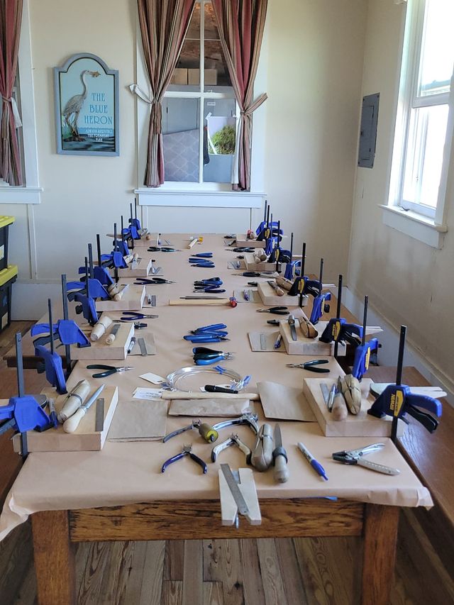 Long wooden table set up for a group jewelry-making workshop with individual bench blocks, blue clamps, pliers, hammers, files and wire coils arranged at each station in a bright room with windows and striped curtains