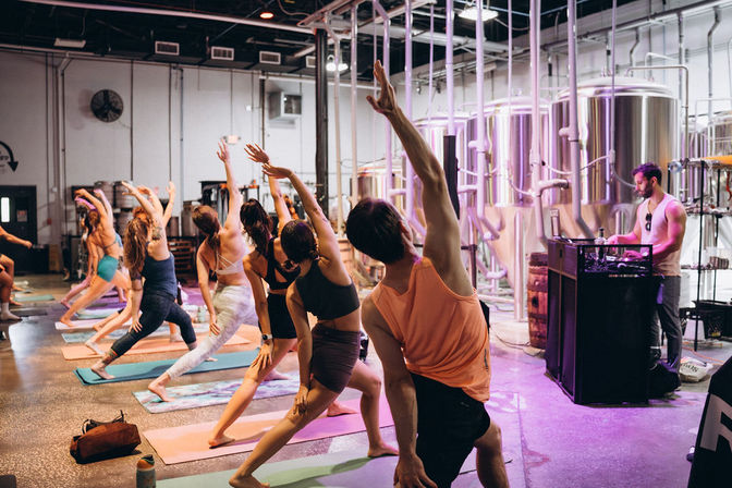 Group yoga class in an urban brewery — people on mats doing side-angle stretches beside stainless steel fermentation tanks while a DJ plays music