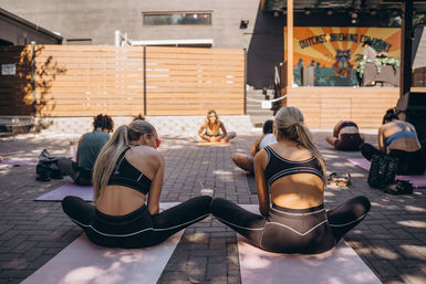 Two women in matching black activewear stretch on pink mats during a sunlit outdoor patio yoga class in an urban courtyard with a colorful mural in the background.