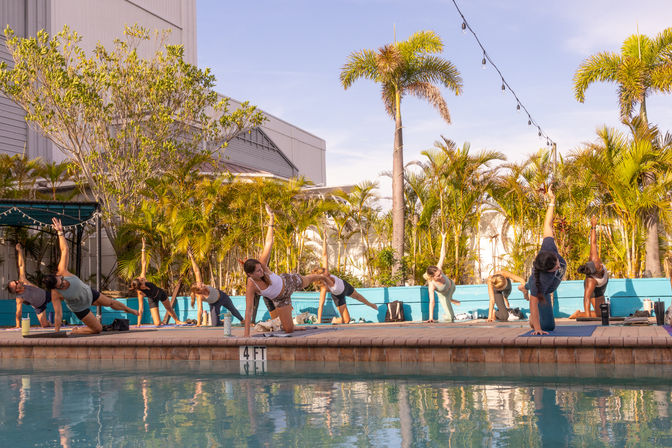 Outdoor poolside yoga class on a sunny tropical patio with palm trees and string lights, a group balancing on mats along the pool edge.