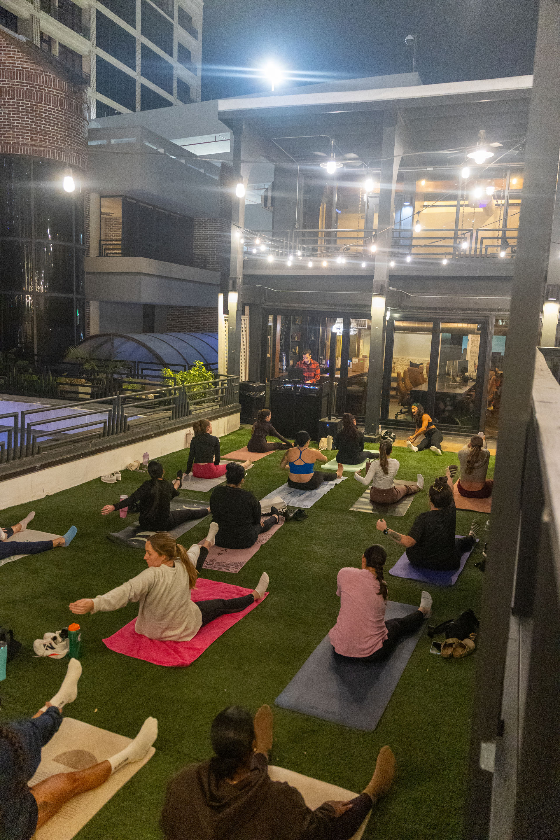 Evening rooftop yoga and stretch class on artificial turf under string lights on an urban rooftop patio, participants on colorful mats stretching while a DJ/instructor plays music by a glass-front building.