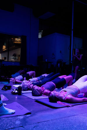 Group of women in bridge pose on yoga mats in a neon-lit indoor fitness studio with purple-blue lighting