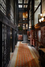 Historic hotel lobby corridor with ornate black wrought-iron elevator cages, patterned mosaic tile runner, red marble wainscoting, decorative wall sconces and a central staircase to a mezzanine