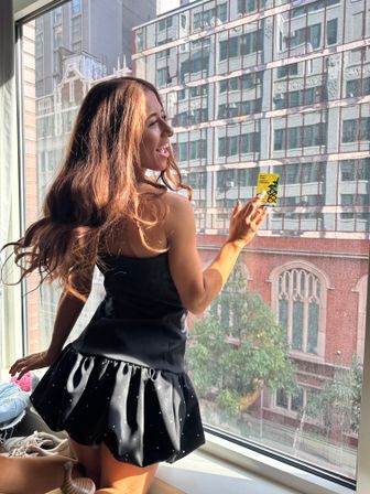 Playful woman in a short black ruffled dress kneeling at a sunlit city apartment window, holding a small yellow sticker against the glass with brick and glass buildings visible outside.