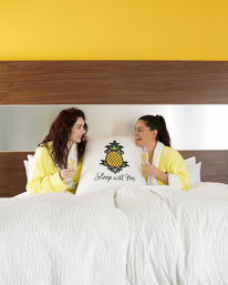Two people in yellow robes laughing on a white bed, clinking champagne flutes around a pillow with a pineapple graphic and the phrase 'Sleep with Me' in a bright modern bedroom.