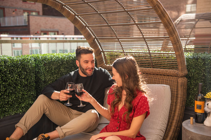 Couple clinking red-wine glasses while relaxing in a wicker daybed on a sunlit urban rooftop balcony with hedge greenery and a bottle of wine nearby.