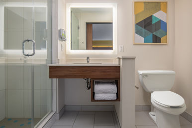 Bright modern bathroom with glass shower, floating wood vanity, LED-lit mirror, neatly folded towels, white toilet and geometric wall art
