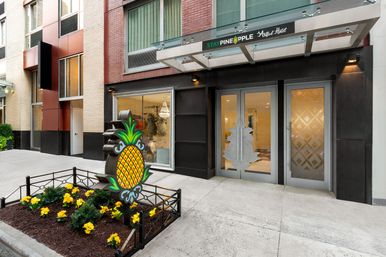 Downtown boutique hotel entrance with a playful pineapple sign, planted bed of yellow flowers, glass double doors and a modern glass awning on a brick facade.