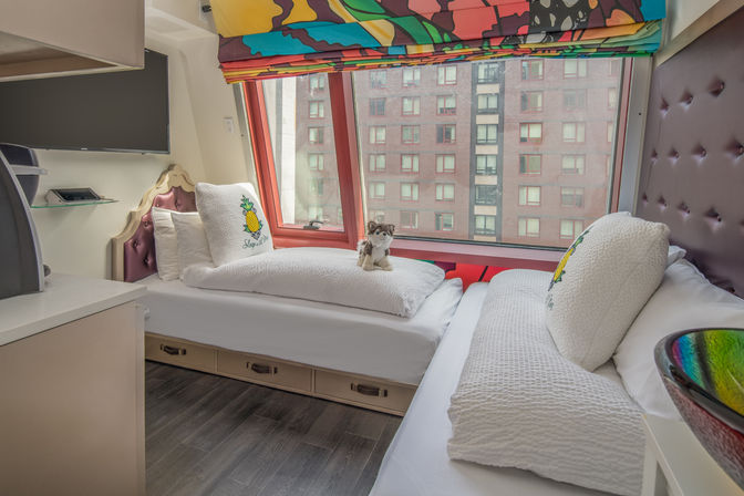 Cozy urban hotel room with two white twin daybeds under a large window facing brick apartment buildings, colorful patterned valance, tufted headboards, pineapple-accented pillows, a small plush toy and under-bed storage drawers.