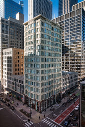 Ornate historic mid-rise on a downtown corner surrounded by glass skyscrapers, crosswalks, cars and a red bus lane.