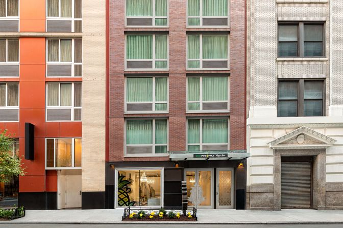 Urban building façade with red brick and beige masonry, rows of green-curtained windows, ground-level glass storefront featuring a pineapple mural, entrance canopy and sidewalk planters on a city street.