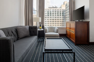 Modern downtown hotel living area with gray tufted sofa and armchair, striped carpet, black metal coffee table, wooden TV cabinet and lamp, large window framing city office buildings and urban view.