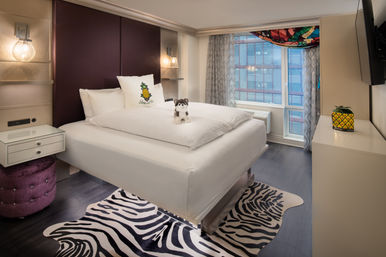 Bright modern downtown hotel room with elevated white bed, pineapple accent pillow and small plush husky on the duvet, zebra-print rug, purple tufted ottoman, and large city-view window with a colorful valance.