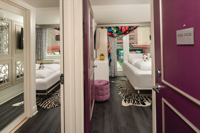 Open purple hotel room door revealing a trendy urban guestroom with white beds, zebra-print rugs, dark wood floors, a purple tufted ottoman, colorful drapery, playful stuffed animals on the beds and a small balcony seating area with a city view.