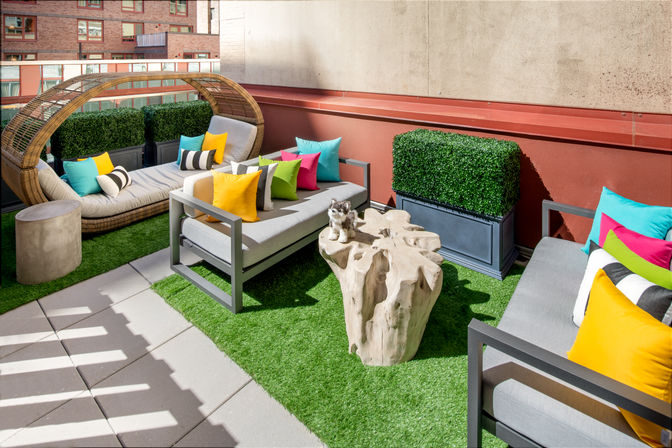 Urban rooftop patio with modern gray sofas, wicker daybed, colorful throw pillows, tree-stump coffee table and boxwood planters on artificial turf.